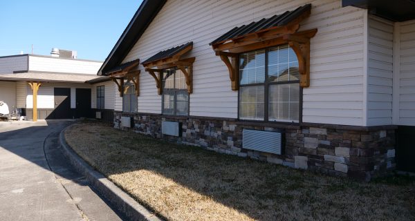 Single-story building with beige siding, stone base, wooden window awnings, driveway and dry lawn