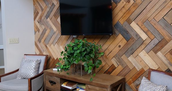 Wall-mounted TV above console table with potted plant and books on herringbone wood accent wall flanked by two armchairs.
