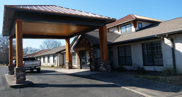Covered entrance with wooden pillars and stone bases at a low-rise building, pickup truck parked under the porte-cochere.