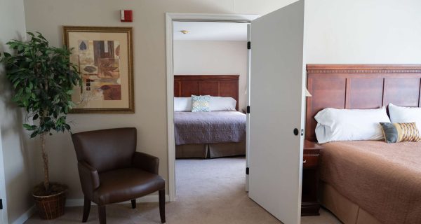 Open doorway between two bedrooms showing brown bedspread, wooden headboard, armchair, potted plant, framed wall art.