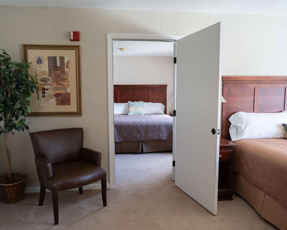 Spacious room with two beds, wooden headboards, open interior door, brown leather chair and potted plant