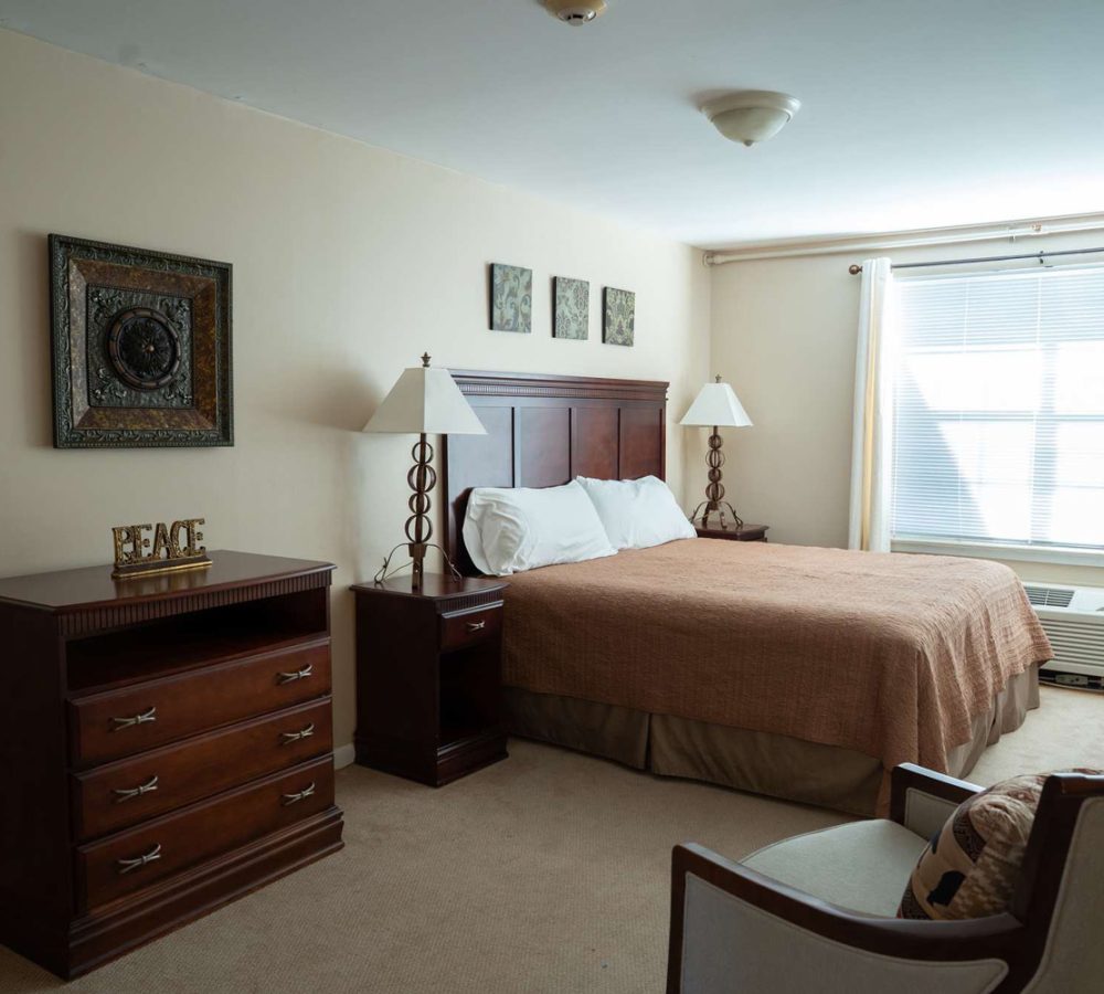 Well style bedroom with queen bed, wooden headboard, matching nightstands and dresser, window with blinds.