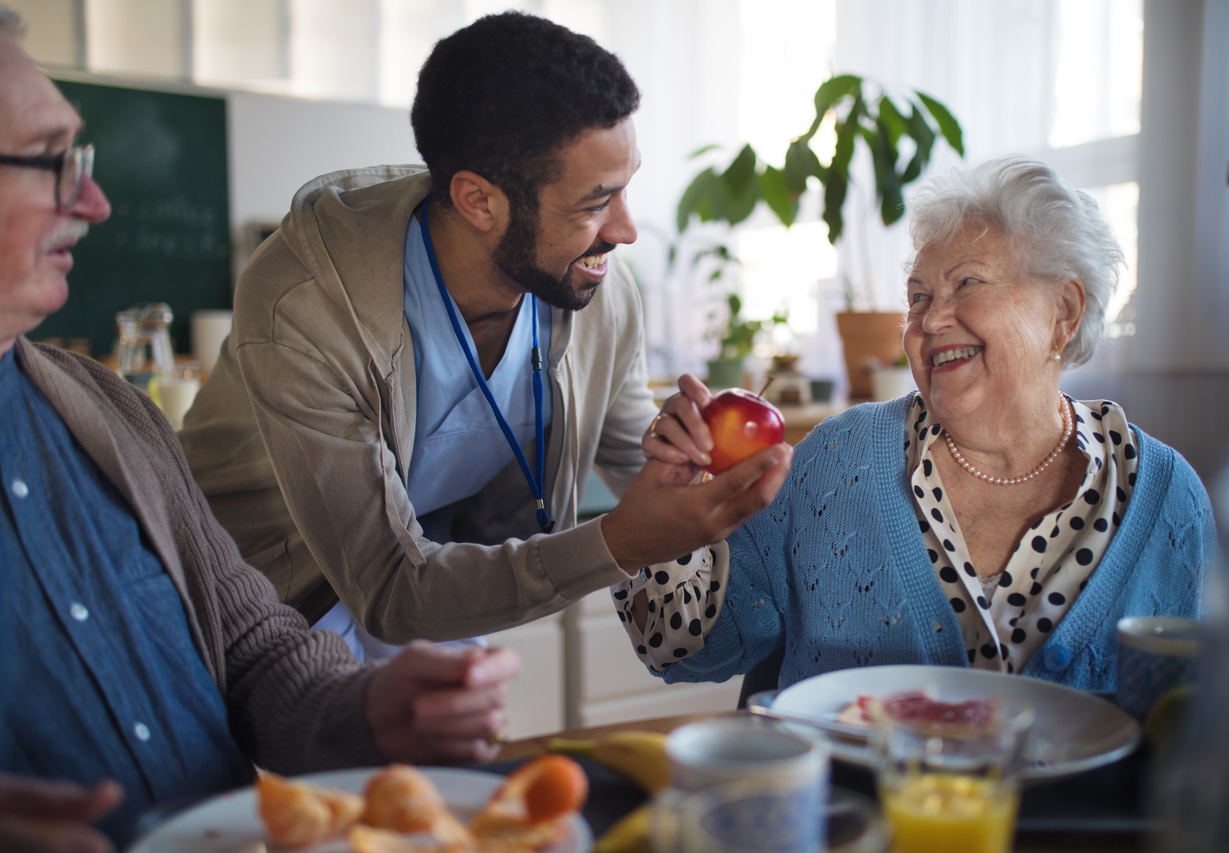 Caregiver handing an apple to a smiling elderly woman at a communal breakfast table in assisted living.