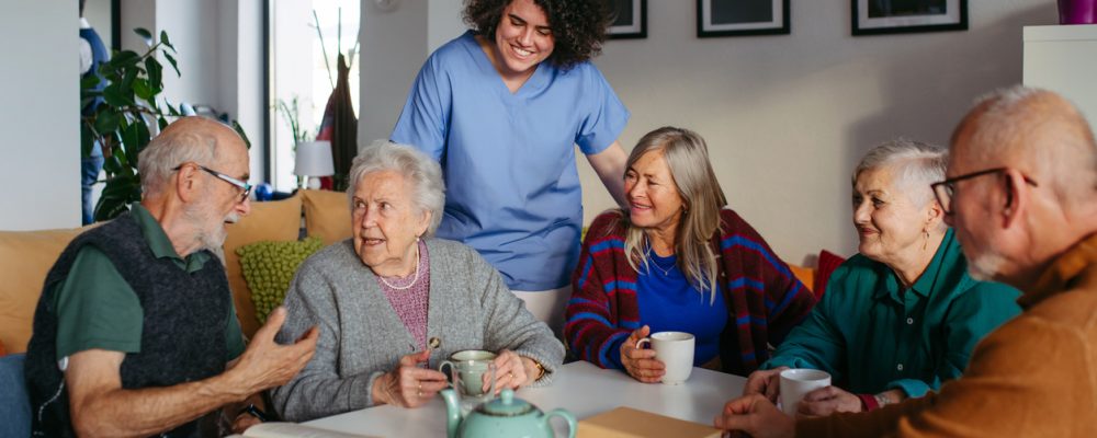 Assisted living caregiver in blue scrubs smiles as a group of seniors chat over tea around a table in a cozy common room