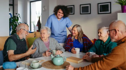 Assisted living caregiver in blue scrubs smiles as a group of seniors chat over tea around a table in a cozy common room