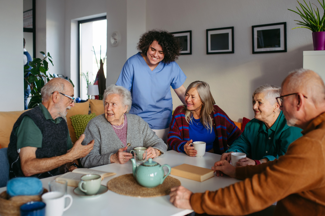Assisted living caregiver in blue scrubs smiles as a group of seniors chat over tea around a table in a cozy common room