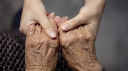 Younger hands gently clasping an aging parent's hands in a moment of support and reassurance