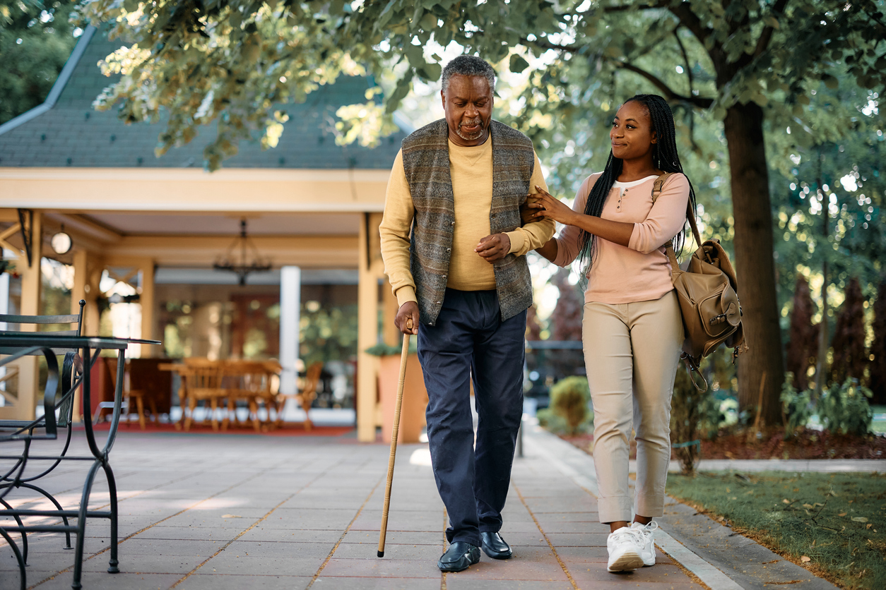 A young woman helps her elderly father walk with a cane along a tree-lined path outside an assisted living community