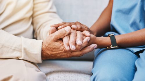 Person in blue scrubs holding an elderly person's hands in a comforting gesture on a couch