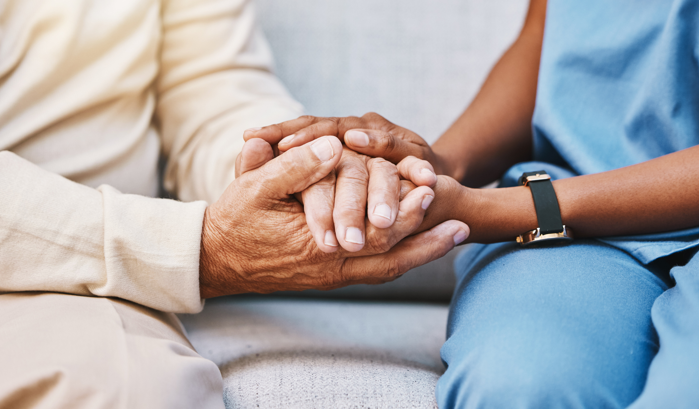 Person in blue scrubs holding an elderly person's hands in a comforting gesture on a couch