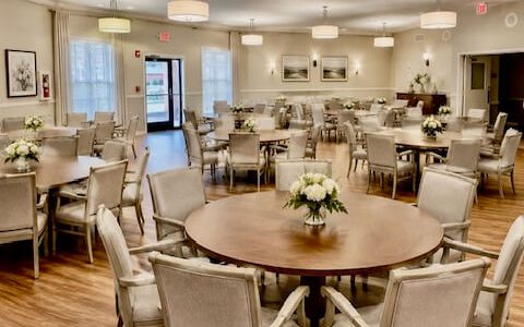 Spacious dining hall with round wooden tables, upholstered chairs, pendant lights and floral centerpieces on wood floors