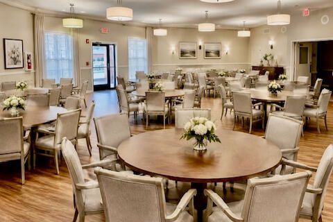 Spacious dining hall with round wooden tables, upholstered chairs, pendant lights and floral centerpieces on wood floors