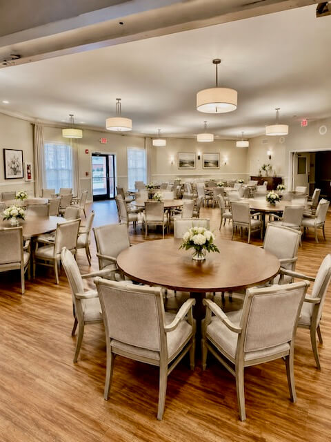 Spacious dining hall with round wooden tables, upholstered chairs, pendant lights and floral centerpieces on wood floors