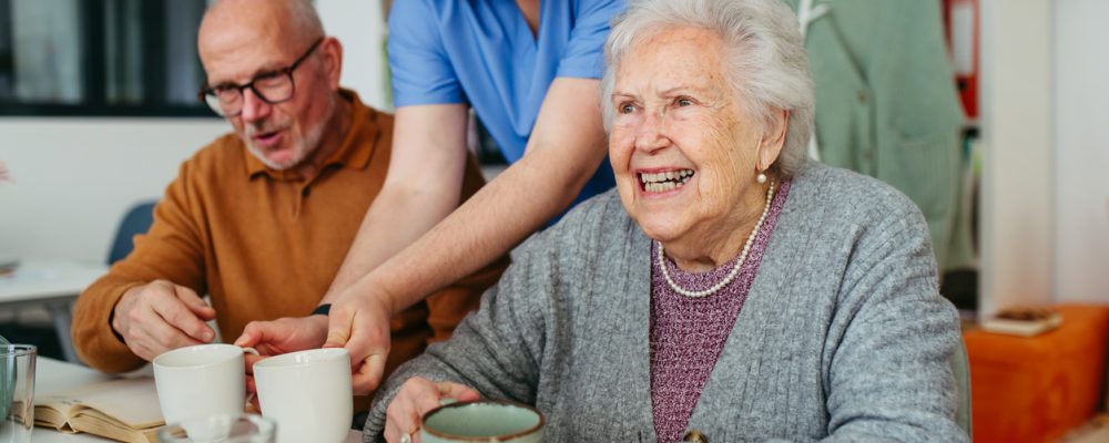Caregiver serving tea to two smiling seniors at a table in a small assisted living home, showing personalized senior care.