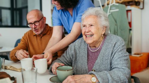 Caregiver serving tea to two smiling seniors at a table in a small assisted living home, showing personalized senior care.
