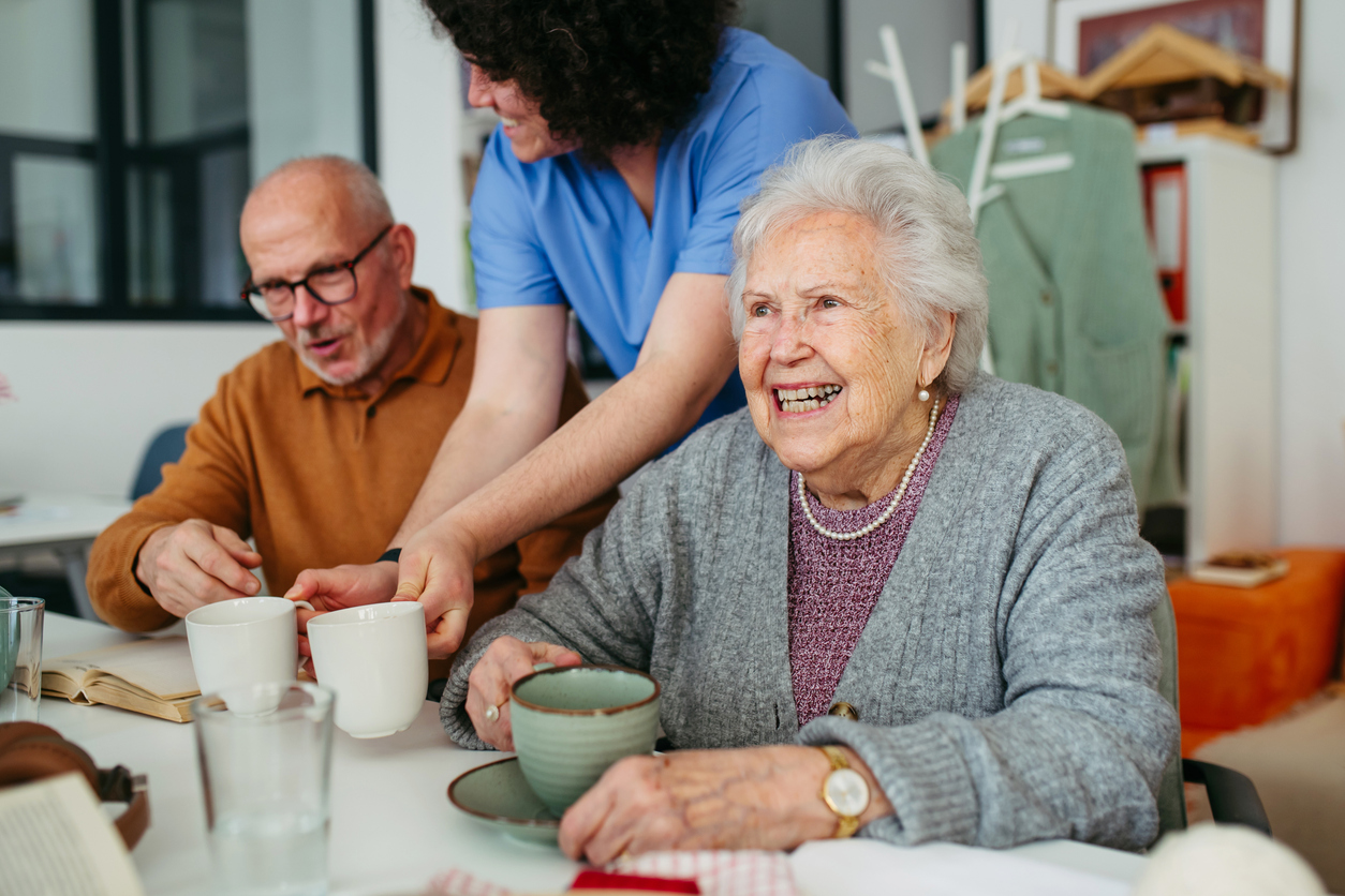 Caregiver serving tea to two smiling seniors at a table in a small assisted living home, showing personalized senior care.
